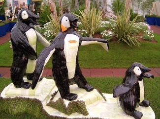 The image features three sculptures of penguins made from eggplant, leek, carrot, asparagus, olive, red pepper, and pumpkin parts, displayed in a garden setting. One sculpture stands upright on a pedestal, another is behind, and the third is smaller, likely representing a juvenile, positioned at the front of the line. The penguins are black and white with touches of orange around the beak and feet. They are set against a backdrop of lush green plants and flowers, suggesting an outdoor exhibition or garden show.